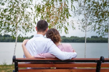 Pembe elbiseli bir adam ve bir kadın kameraya sarılıp nehir kıyısındaki bankta oturuyorlar. Düğün günü, şenlikli bir olay, yeni evlenmiş. Yakın çekim.