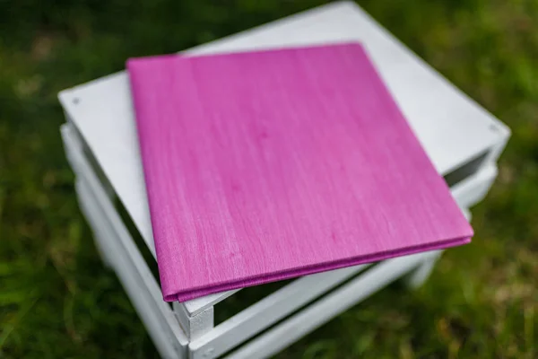 Book, album, notebook or photobook with purple leather cover lying on white wooden box among green grass. Soft focused shot.