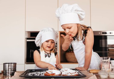 Küçük kızlar mutfakta beyaz aşçı şapkalarıyla oyun oynuyorlar.