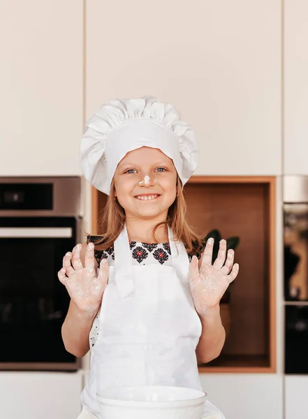 A little funny girl in a white chef hat plays with flour in the kitchen. Baby cooks