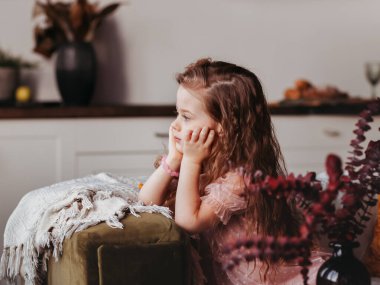Küçük düşünceli bir kız oturma odasındaki kanepede oturur ve pencereden dışarı bakar.
