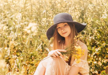 Şapkalı genç ve güzel bir kız tarlanın arka planında bir buket kolza sahip.