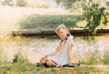 İki örgülü kız yazın nehir kenarında çimlerin üzerinde oturur. Yaz bahçesinde gülümseyen çocuk