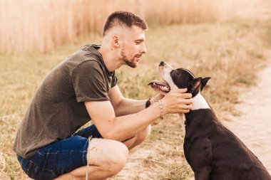 Genç bir adam pitbull terrier cinsinden bir köpekle doğada yürüyor.