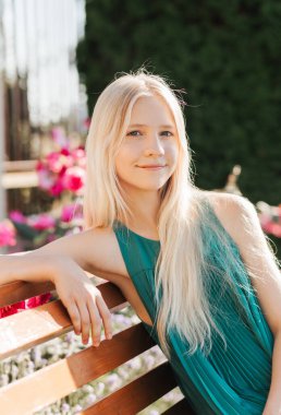 Uzun sarı saçlı güzel bir genç kız çiçek açan bir parktaki bankta oturuyor.