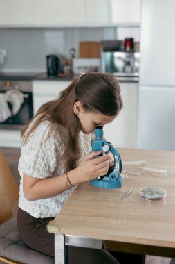 Okul çağındaki kız evde mikroskop kullanarak masada oturuyor, odaklanmış ve meraklı, evde eğitim, fen eğitimi, öğrenim.