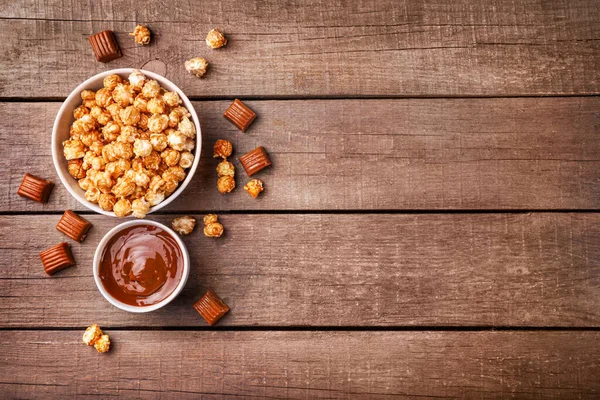 Sweet caramel popcorn and melted caramel on wooden background. Top view. Copy space.