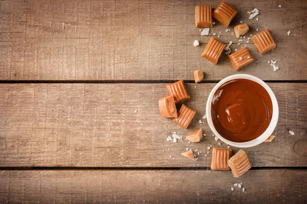 Melted caramel in bowl with salted caramel candies on wooden background. Top view. Copy space.