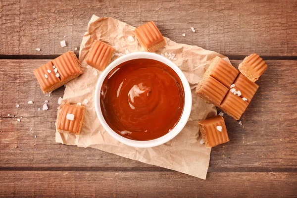 Melted caramel in bowl with salted caramel candies on wooden background. Top view.