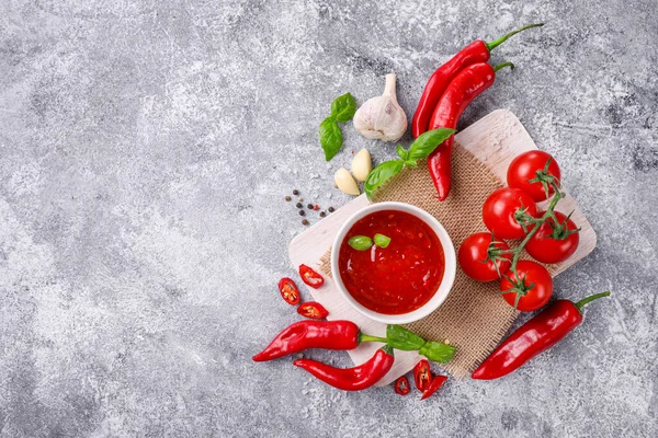 Hot spicy chili sauce in small bowl with red chili pepper and tomatoes on burlap on cutting board on stone background. Top view. Copy space.