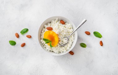 Beyaz kasede taze süzme peynir, nane yaprakları, şeftali parçaları, badem fıstığı ve beyaz arka planda çay kaşığı. Üst görünüm.