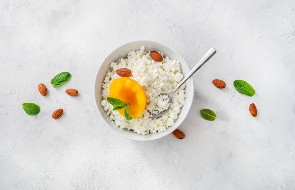 Beyaz kasede taze süzme peynir, nane yaprakları, şeftali parçaları, badem fıstığı ve beyaz arka planda çay kaşığı. Üst görünüm.