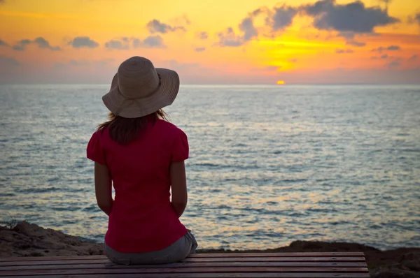 Jeune femme en chapeau assis et regardant le coucher du soleil — Photo
