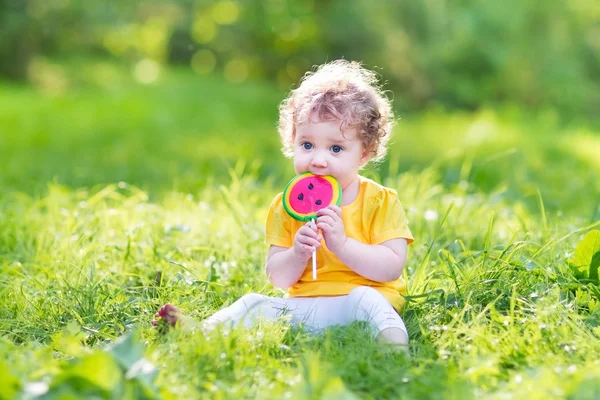 Χαριτωμένο μωρό σγουρά κορίτσι τρώει καρπούζι καραμέλα σε ένα ηλιόλουστο πάρκο — Φωτογραφία Αρχείου