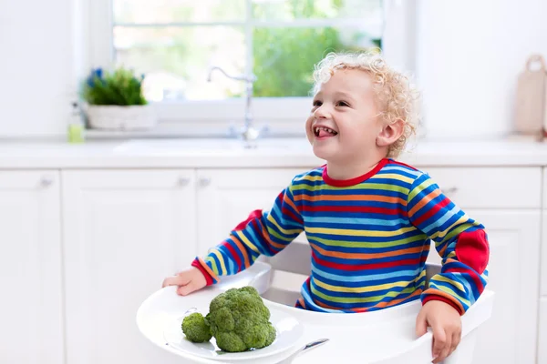 Little boy eating broccoli in white kitchen — Stock Photo, Image