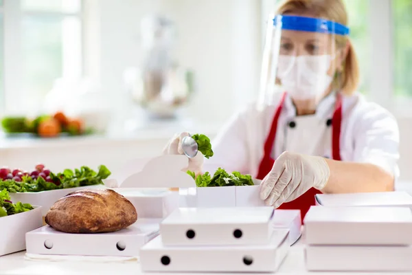 Food takeaway in restaurant in coronavirus outbreak. Cafe staff in face mask and shield cooking takeout order. Safety, covid-19 prevention. Chef with salad in cardboard container, eco friendly, vegan.