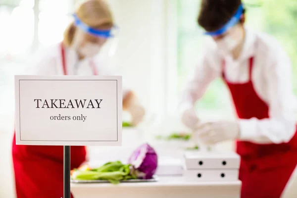 Food takeaway in restaurant in coronavirus outbreak. Cafe staff in face mask and shield cooking takeout order. Safety, covid-19 prevention. Chef with salad in cardboard container, eco friendly, vegan.