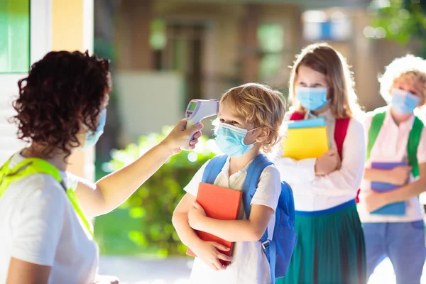 Temperature screening and medical check at school. Child in face mask in class in covid-19 outbreak. Teacher with thermometer at preschool entrance. Social distancing. Coronavirus prevention.