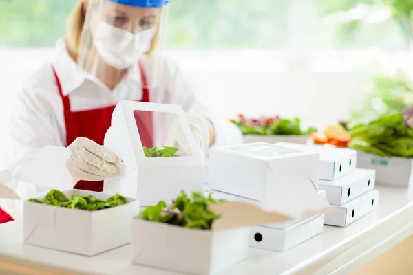 Food takeaway in restaurant in coronavirus outbreak. Cafe staff in face mask and shield cooking takeout order. Safety, covid-19 prevention. Chef with salad in cardboard container, eco friendly, vegan.