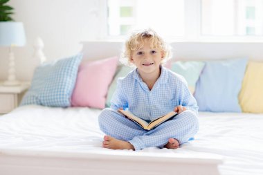 Beyaz güneşli yatak odasında pencereli çocuk kitapları okuyor. Çocuklar kitap okur. Çocuk odası. Evde pijamalı sarışın kıvırcık çocuk. Bebek odası için yatak ve uyku giysileri. Oyuncaklı çocuk.