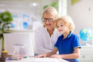 Çevrimiçi uzaktan öğrenme. Bilgisayarlı okul çocukları öğretmen ve sınıf grubuyla görüntülü konuşma yapıyorlar. Anne oğluna yardım ediyor. Çocuk evden ders çalışıyor. Koronavirüs salgınında evde eğitim.