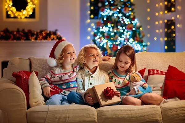 クリスマスツリーの子供たちとクリスマスの前夜の暖炉 自宅でクリスマスを祝う子供たちと家族 白いソファの開口部のプレゼントにニットセーターの男の子と女の子 子供のための休日の贈り物 — ストック写真