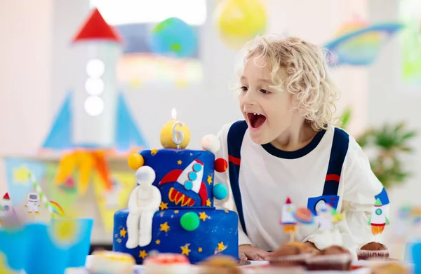 Kids space theme birthday party with cake and cupcakes. Rocket, solar system planet and spaceship decoration for child event. Little boy in astronaut costume blowing candles and opening presents.