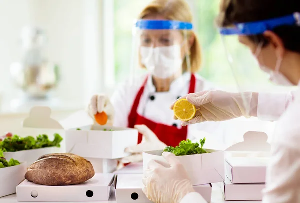 Food takeaway in restaurant in coronavirus outbreak. Cafe staff in face mask and shield cooking takeout order. Safety, covid-19 prevention. Chef with salad in cardboard container, eco friendly, vegan.