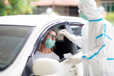 Coronavirus geniz temizleme testi. Tehlikeli madde giysisi giymiş bir doktor, salya örneği alıp Coperid-19 teşhisi için arabada test yapıyor. Hastanedeki sürücü kadın hasta test istasyonunda. Virüs salgını.