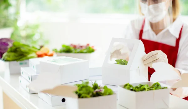 Food takeaway in restaurant in coronavirus outbreak. Cafe staff in face mask and shield cooking takeout order. Safety, covid-19 prevention. Chef with salad in cardboard container, eco friendly, vegan.