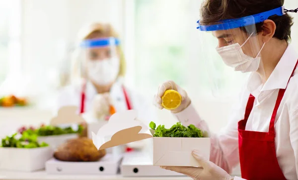 Food takeaway in restaurant in coronavirus outbreak. Cafe staff in face mask and shield cooking takeout order. Safety, covid-19 prevention. Chef with salad in cardboard container, eco friendly, vegan.