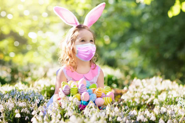 在Covid 19疫情中 戴着面具的小女孩在寻找复活节彩蛋上玩得很开心 穿着兔子耳朵和兔子服装的孩子们在庆祝科罗纳威斯大流行 有彩蛋的孩子安全庆祝活动 — 图库照片