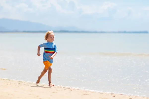 熱帯のビーチで遊んでいる子供たち 子供たちは夏休みに海で泳いだり遊んだりします 砂と水の楽しさ 幼い子供のための太陽の保護 海の岸を走って飛び跳ねる少年 — ストック写真