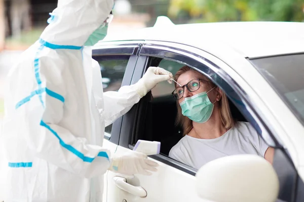 Coronavirus nasal drive-through swab test. Doctor in hazmat suit taking saliva sample for covid-19 diagnostics in a car. Woman driver patient at drive-thru testing station in hospital. Virus outbreak.