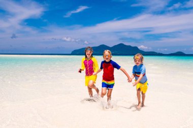 Tropik plajda oynayan çocuklar. Çocuklar yaz tatilinde yüzer ve denizde oynarlar. Kum ve su eğlencesi, küçük çocuklar için güneş koruması. Okyanus kıyısında koşan ve zıplayan küçük çocuk.