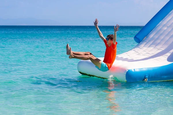 Kids jumping on trampoline on tropical sea beach. Children jump on inflatable water slide. Aqua amusement park in exotic island resort. Family vacation and travel with child. Ocean coast fun.