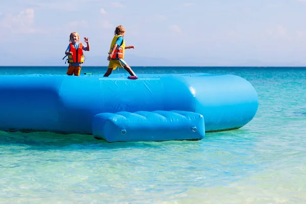 Kids jumping on trampoline on tropical sea beach. Children jump on inflatable water slide. Aqua amusement park in exotic island resort. Family vacation and travel with child. Ocean coast fun.