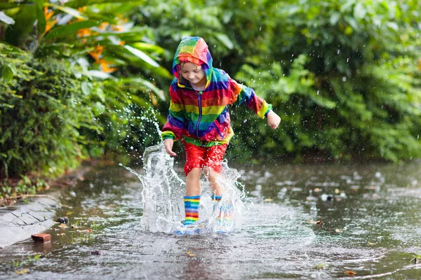 Kid playing in the rain in autumn park. Child jumping in muddy puddle on rainy fall day. Little boy in rain boots and rainbow jacket outdoors in heavy shower. Kids waterproof footwear and coat.