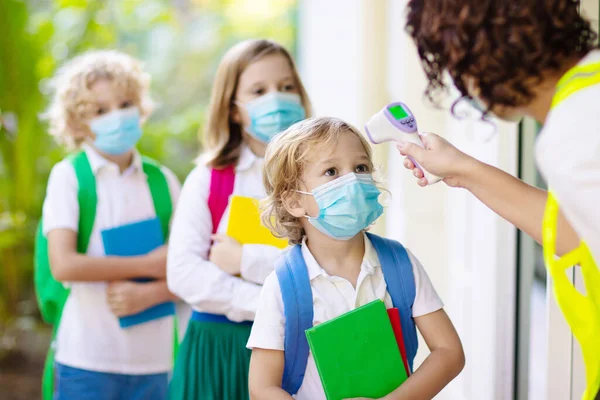 Temperature screening and medical check at school. Child in face mask in class in covid-19 outbreak. Teacher with thermometer at preschool entrance. Social distancing. Coronavirus prevention.