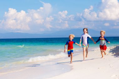 Tropik plajda oynayan çocuklar. Çocuklar yaz tatilinde yüzer ve denizde oynarlar. Kum ve su eğlencesi, küçük çocuklar için güneş koruması. Küçük çocuk ve kız okyanus kıyısında koşup zıplıyorlar..