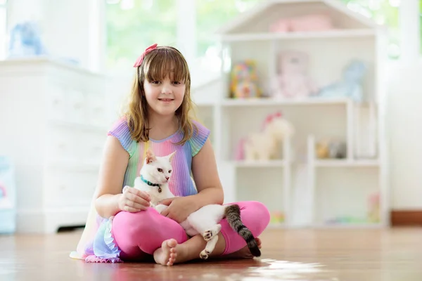 Child playing with cat at home. Kids and pets. Little girl feeding and petting cute white cat. Cats tree and scratcher in living room interior. Children play and feed kitten. Home animals.