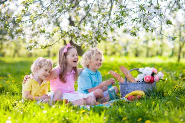 Bahar Parkı 'nda çiçek açan kiraz ağaçlarıyla aile pikniği. Çocuklar dışarıda meyve ve ekmek yiyorlar. Çiçekli elma bahçesinde piknik sepetiyle battaniyenin üzerinde oturuyorlar. Çocuklar için sağlıklı beslenme.
