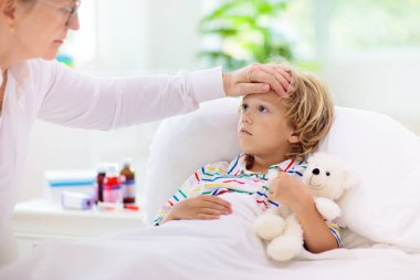 Hasta küçük çocuk ilaçla. Anne yatakta hasta çocuk ateşini kontrol ediyor. Astım tedavisi için astım spreyi olan hasta bir çocuk. Grip mevsimi. Ebeveyn yatak odasında ya da genç hastalar için hastane odasında.