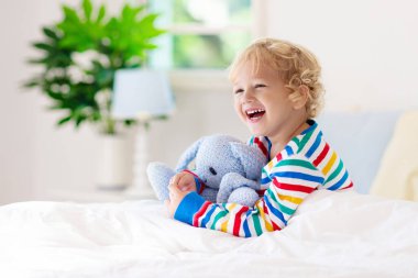 Beyaz güneşli bir odada pencereli bir çocuk yatakta oynuyor. Çocuk odası ve iç tasarım. Erkek bebek evde. Çocuk yuvası için yatak ve tekstil. Oyuncağı ve kitabı olan bir çocuk. Uyku ve uyku zamanı..