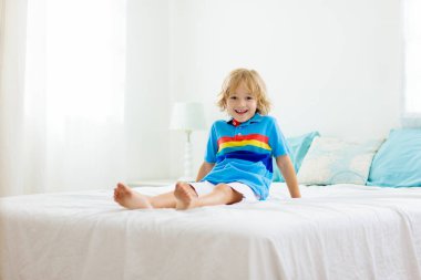 Beyaz güneşli bir odada pencereli bir çocuk yatakta oynuyor. Çocuk odası ve iç tasarım. Erkek bebek evde. Çocuk yuvası için yatak ve tekstil. Oyuncağı ve kitabı olan bir çocuk. Uyku ve uyku zamanı..