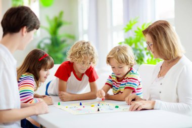 Aile evde masa oyunu oynuyor. Çocuklar stratejik oyunlar oynar. Küçük çocuk zar atıyor. Yaz tatili için eğlenceli bir ev aktivitesi. Kardeşler arası bağ. Eğitici oyuncaklar. Arkadaşlar oyun gecesinin tadını çıkarın.