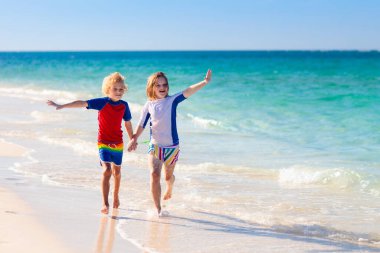 Tropik plajda oynayan çocuklar. Çocuklar yaz tatilinde yüzer ve denizde oynarlar. Kum ve su eğlencesi, küçük çocuklar için güneş koruması. Küçük çocuk ve kız okyanus kıyısında koşup zıplıyorlar..