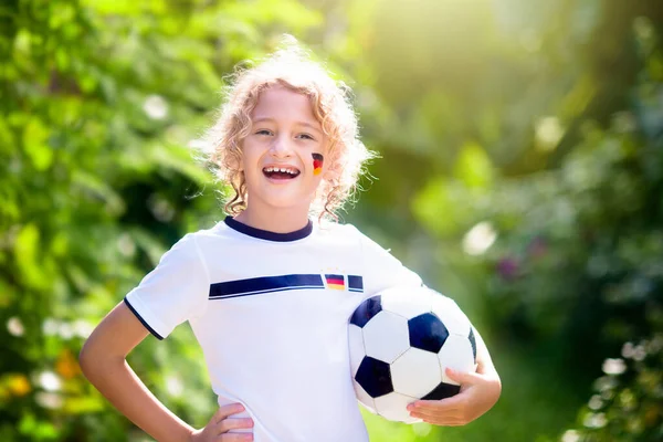 Kids play football on outdoor field. Germany team fans with national flag. Children score a goal at soccer game. Child in German jersey and cleats kicking ball. Fan celebrating victory at pitch.