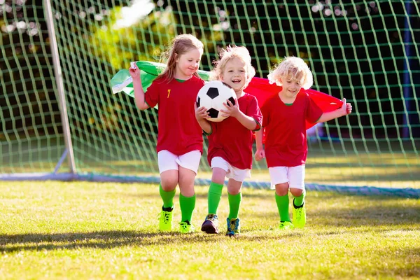 Kids play football on outdoor field. Portugal team fans with national flag. Children score a goal at soccer game. Child in Portuguese jersey and cleats kicking ball. Fan celebrating victory at pitch.