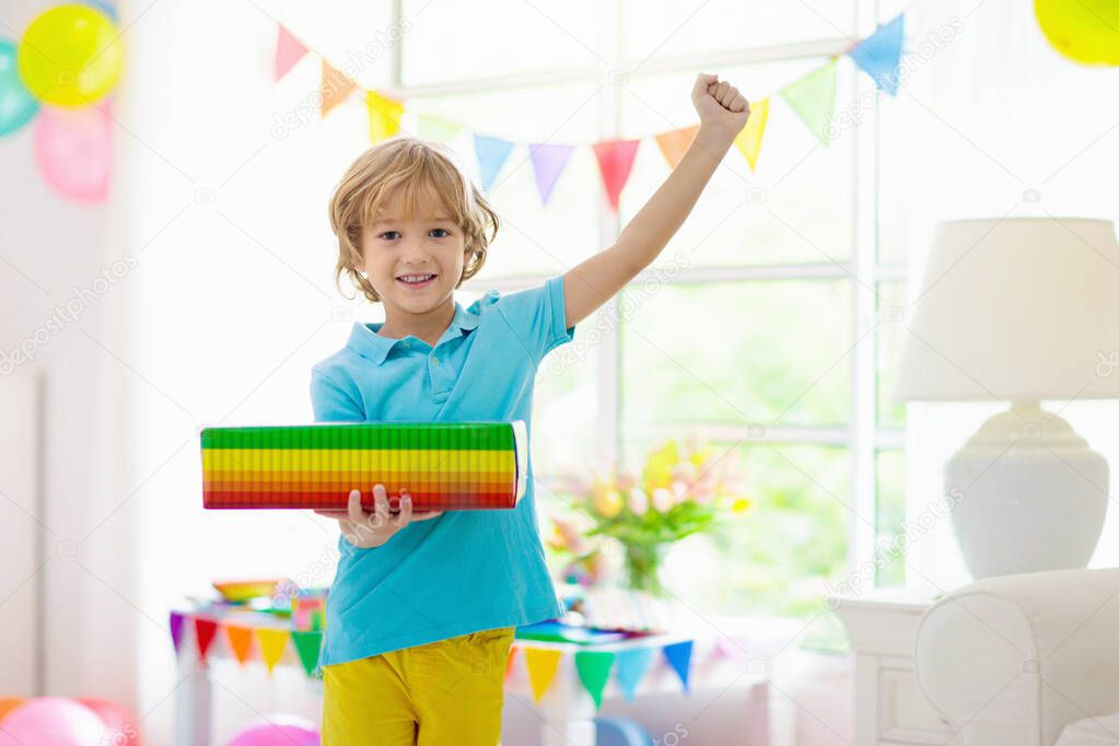 Kids birthday party. Child blowing candles on cake and opening presents. Pastel rainbow theme celebration. Family celebrating at home. Boy opening gifts, eating cakes. Sweets for children.